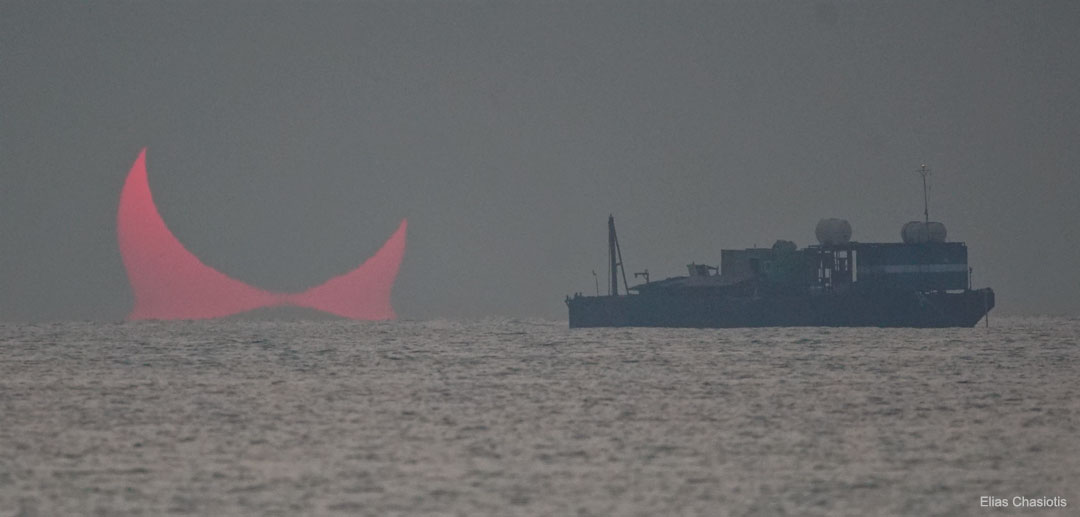 A partially eclipse of a Sun rising over water is shown. 
A ship appears on the right. The Sun appears reddened by the
Intervening Earth’s atmosphere. An inversion layer in the
atmosphere makes part of the Sun appeared doubled near the 
horizon. 
Please see the explanation for more detailed information.