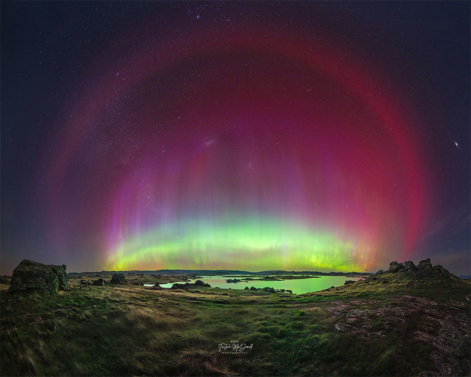 A flat landscape with a pond is imaged at night below
a starfield. A multicoloured aurora is seen in an arc across
 the image centre. Around this arc is another red arc that
is particularly smooth. 
Please see the explanation for more detailed information.