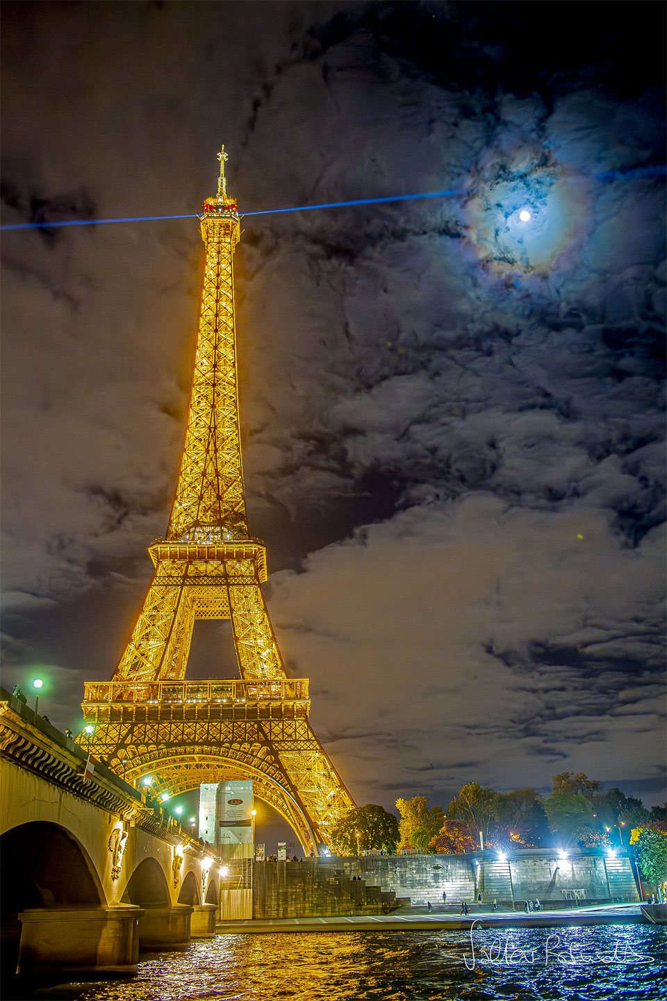 The famous Eiffel Tower in Paris, France is pictured 
on the left lit up in gold at night. A blue laser shines
out from the top. Clouds dot the background sky. The Moon
is also visible through the clouds, but is circled by 
colourful rings: a lunar corona.
Please see the explanation for more detailed information.