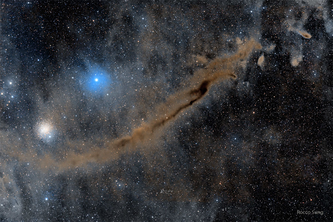A busy starfield is shown which an elongated brown 
nebula running diagonally from the lower left to the 
upper right. A bright blue star and a star cluster 
appear above the nebula. 
Please see the explanation for more detailed information.