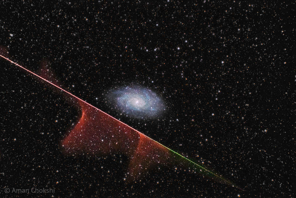 A distant spiral galaxy is seen in the image centre.
A multi-coloured streak runs diagonally across the image
from the upper left to the lower right. Parts of this
streak have gas near it. The background is dark field
filled with stars. 
Please see the explanation for more detailed information.