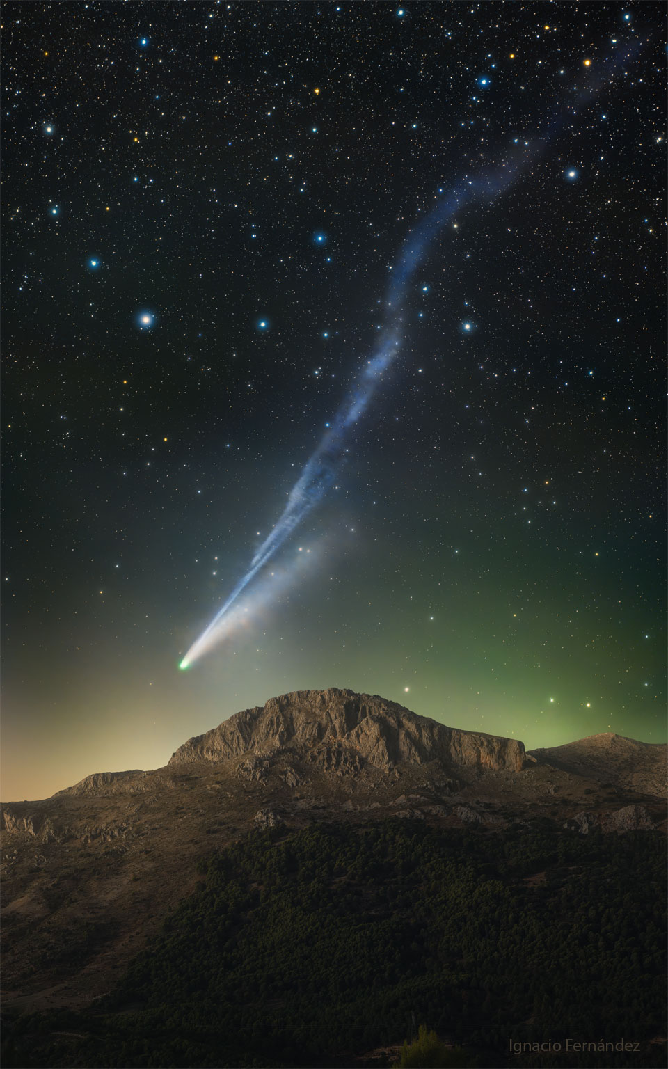 A starfield is shown above a mountain peak. Just
above the mountain and extending up toward the upper 
right is a blue-tinted tail of a comet. The comet's
head is just to the left of the peak.  
Please see the explanation for more detailed information.