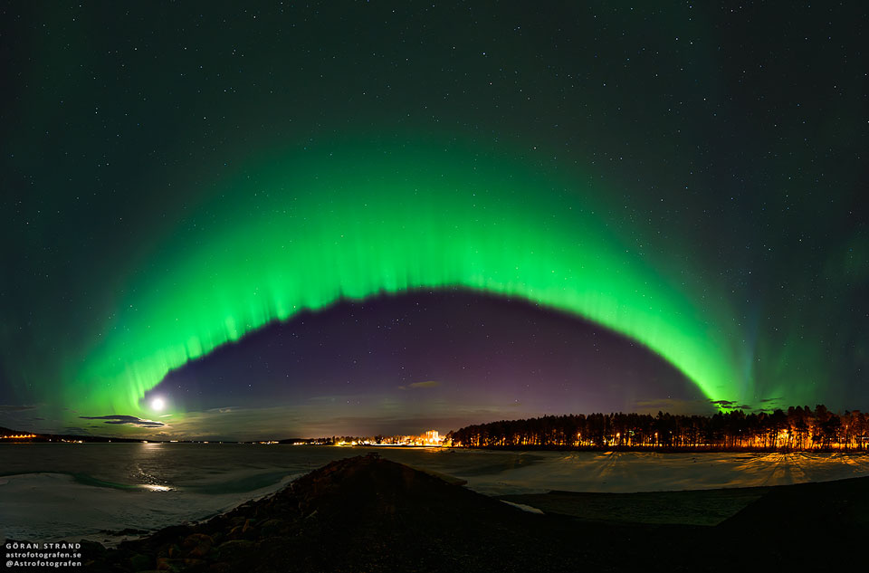 
A big green arc is seen arching across the night sky. The arc
fades away above into a green haze, while no green glow is seen
below the arc. A dark sky filled with stars and constellations
fills the background. Snow and distant trees line the foreground.
Please see the explanation for more detailed information.