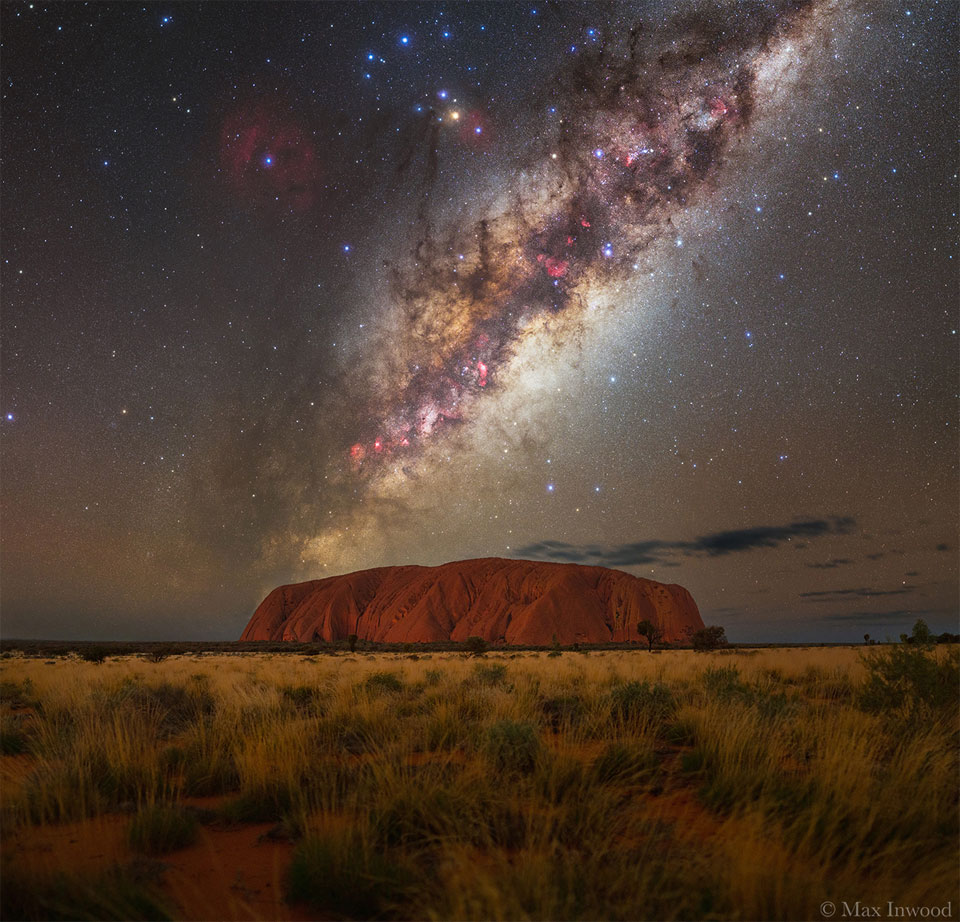 A starry sky is shown with the busy central band of 
our Milky Way Galaxy showing rising diagonally from the lower
right. In the foreground are flat grasslands leading up
to a huge orange rock mound named Uluru.
Please see the explanation for more detailed information.