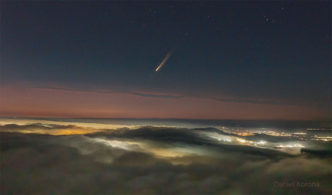 A picture shows clouds across the bottom and a dark night sky
across the top. In the middle is a band of orange sky. City lights
are visible on the right through gaps in the clouds. In the centre
of the upper sky is a comet with its tail pointing toward the 
upper right. 
Please see the explanation for more detailed information.