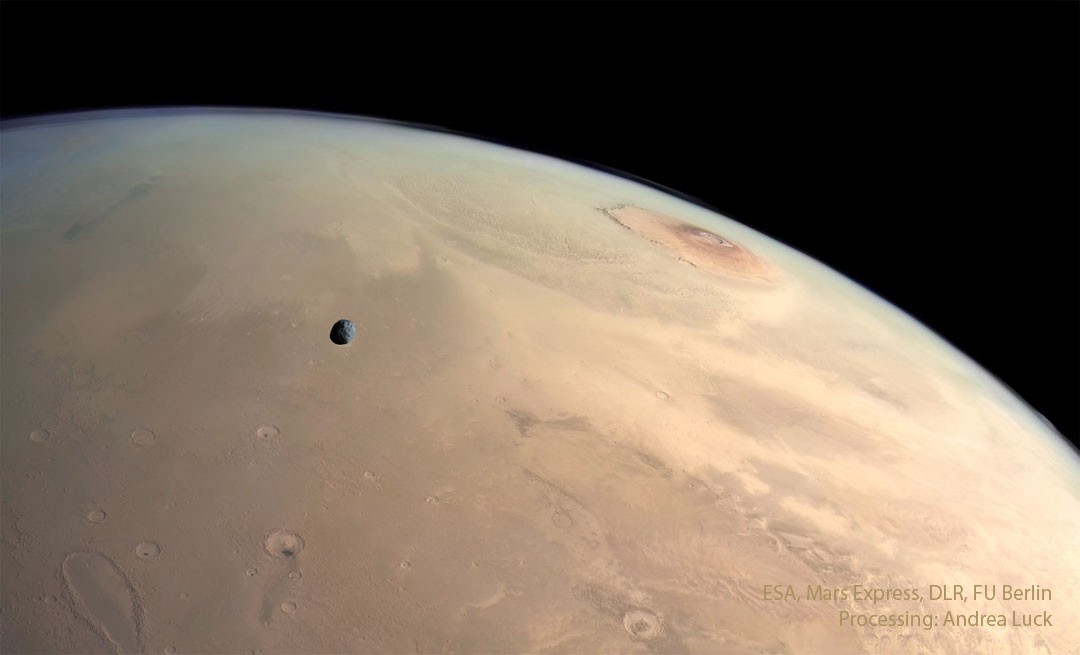 A panoramic view of the surface of Mars. Several landforms are 
visible including craters and volcanos. A small dark moon is superposed
in front of part of the surface. 
Please see the explanation for more detailed information.