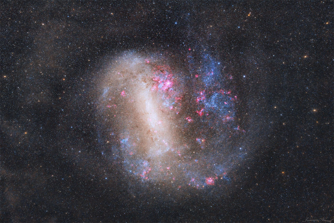 An unusual looking galaxy is shown with a light bar
running nearly vertical and blue stars and red nebulae 
around the edges.  
Please see the explanation for more detailed information.