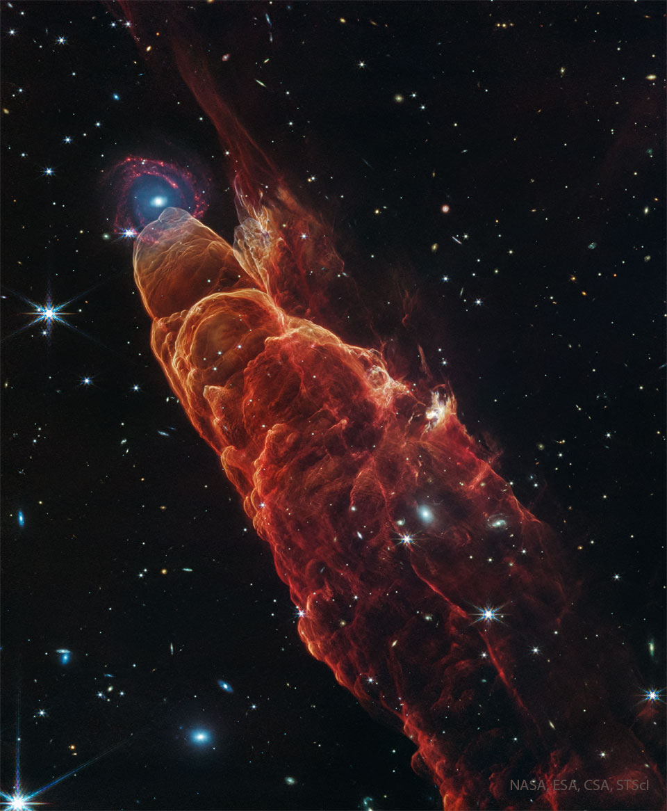 A dark starfield is dominated by a red cone-like
nebula with its base on the bottom right and extending
diagonally through the image. At the peak of the cone
toward the upper left is a background spiral galaxy.
Please see the explanation for more detailed information.