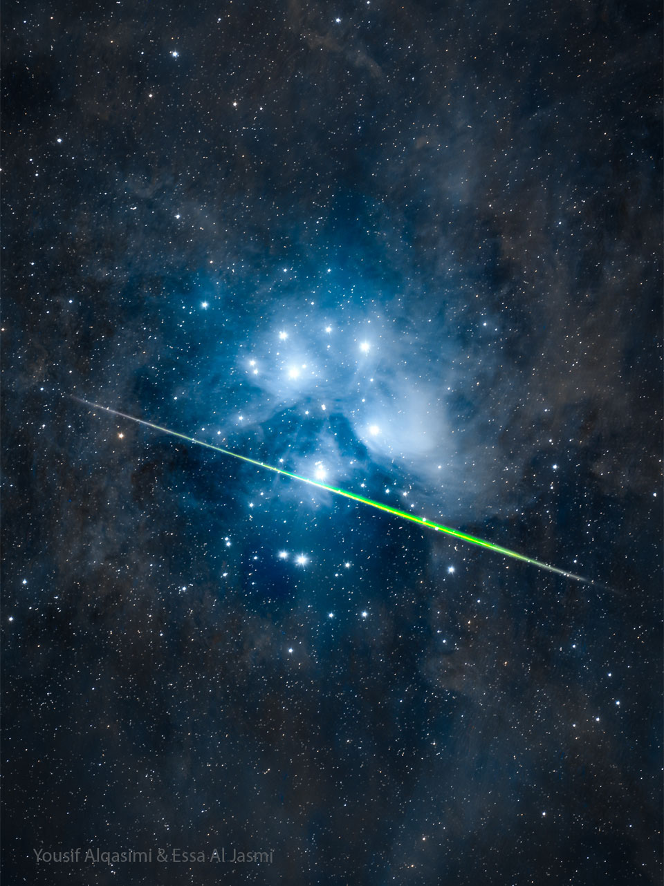A starfield surrounds the bright blue stars of a
star cluster: the Pleiades star cluster. Nearly horizontally
across the cluster is a bright green streak, most likely
a meteor. 
Please see the explanation for more detailed information.