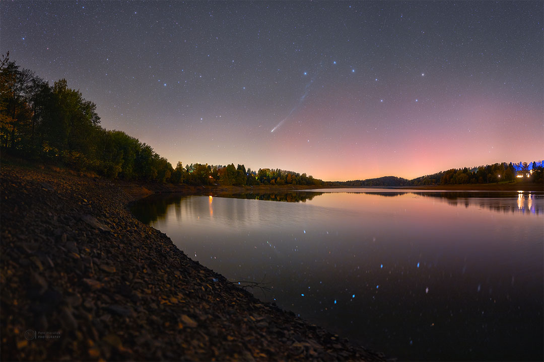 A night sky is seen over a lake. The sky contains
numerous stars including the Big Dipper asterism. Prominent
in the sky is a comet with a long tail. Both the Big Dipper
and the comet are seen reflected in the still water of the 
lake.
Please see the explanation for more detailed information.