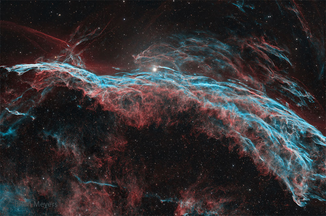A starfield is shown that has a multi-filament
nebula flowing across it horizontally. The most 
prominent colours are red and blue. 
Please see the explanation for more detailed information.