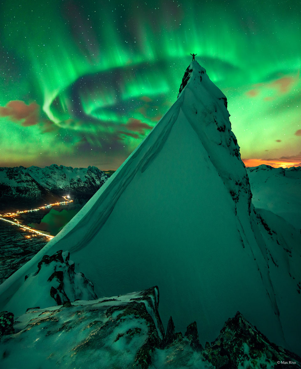 A person with the arms raised stands atop of a
rock peak covered in snow. Snow covered mountains
are all around. Green aurora swirl overhead and 
reflect off the snow. 
Please see the explanation for more detailed information.